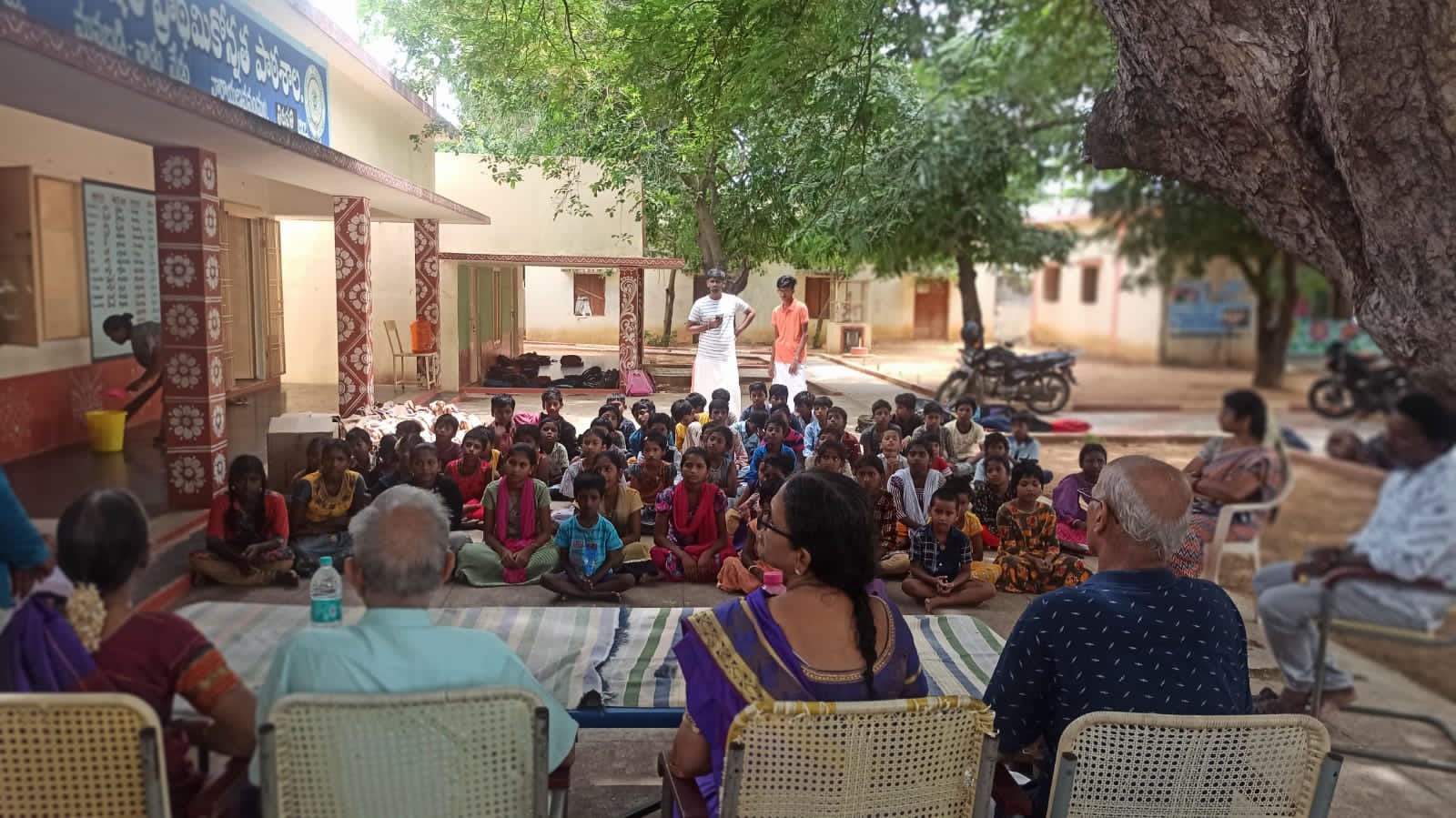 A large group of schoolchildren gathered outdoors with adults seated in front